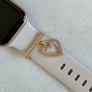 Könnte beinhalten: Ein weißes Apple Watch Armband mit einem goldfarbenen Herz-Anhänger. Der Anhänger ist mit rosa Strasssteinen verziert.