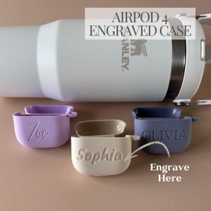 Op de afbeelding: Drie Airpod 4 hoesjes met verschillende gegraveerde namen. De hoesjes zijn paars, beige en blauw. De tekst "Graveer hier" staat op de afbeelding met een pijl die naar het beige hoesje wijst.