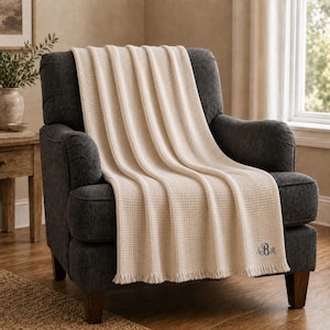 Op de afbeelding: Een crèmekleurige wafelgeweven sprei over een donkergrijze fauteuil. De sprei heeft een gefranjerde rand en geborduurde initialen in de hoek. De stoel staat in een goed verlichte kamer met een raam en een bijzettafel.