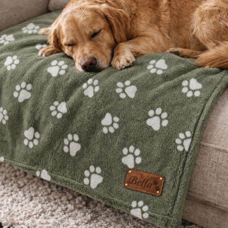 May include: A soft olive green fleece blanket featuring a repeating pattern of white paw prints. A rectangular brown leather-like patch is stitched to the bottom corner, displaying the name Bella in cursive script with a small paw print icon.