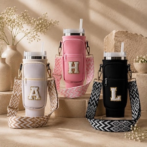 Peut inclure: Trois porte-gobelets isothermes en blanc, rose et noir, chacun doté d'une poche frontale zippée, d'une lettre en relief à bordure dorée et d'une bandoulière à motifs amovible. Chaque étui contient un gobelet en acier inoxydable avec une paille.