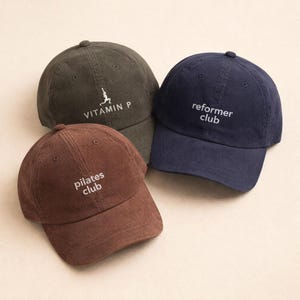 May include: Three corduroy baseball caps in olive green, navy blue, and brown. The olive green cap has the text "VITAMIN P" and a graphic. The navy blue cap has the text "reformer club". The brown cap has the text "pilates club".