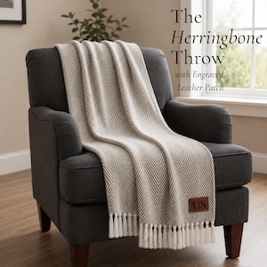 May include: A beige and white herringbone patterned throw blanket with white fringe tassels at the bottom edge. A brown rectangular leather patch is attached to the lower corner, featuring the engraved letters LS. Text reads: The Herringbone Throw with Engraved Leather Patch.