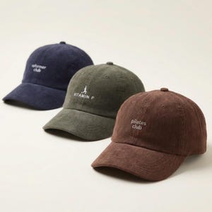 May include: Three corduroy baseball caps in navy blue, olive green, and brown. Each cap has a curved brim and a small embroidered logo: "reformer club", "VITAMIN P", and "pilates club". The caps are arranged in a row.