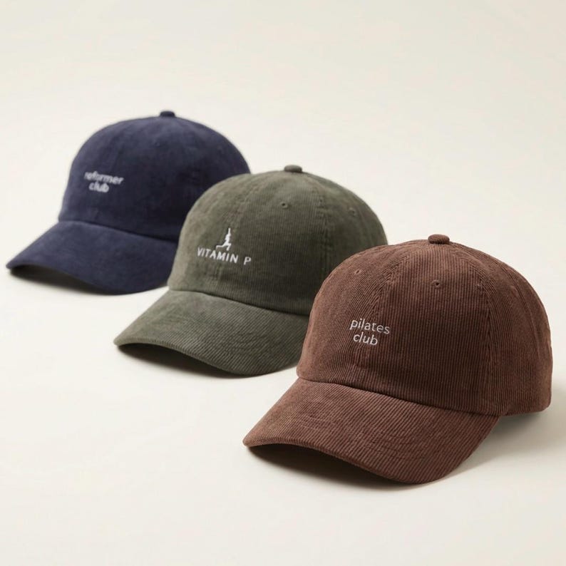 May include: Three corduroy baseball caps in navy blue, olive green, and brown. Each cap has a curved brim and a small embroidered logo: "reformer club", "VITAMIN P", and "pilates club". The caps are arranged in a row.