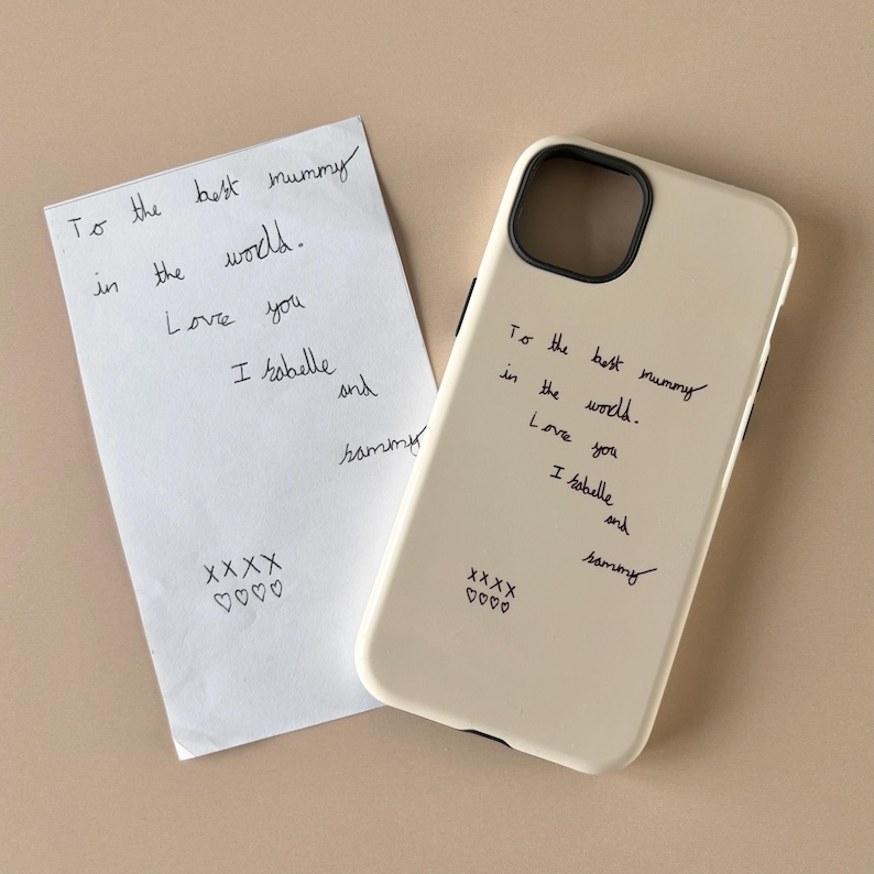 Może przedstawiać: Kremowe etui na telefon z czarnym, odręcznym napisem: To the best mummy in the world. Love you Isobelle and Sammy, po kt&oacute;rym następują cztery X i cztery serca. Obok znajduje się biała kartka papieru z tą samą odręczną wiadomością.