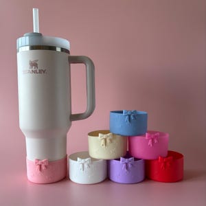 Może przedstawiać: Biały kubek podróżny Stanley z szarym uchwytem i szarą pokrywką. Kubek stoi na różowym tle z siedmioma silikonowymi podkładkami w różnych kolorach, każda z wzorem kokardki.
