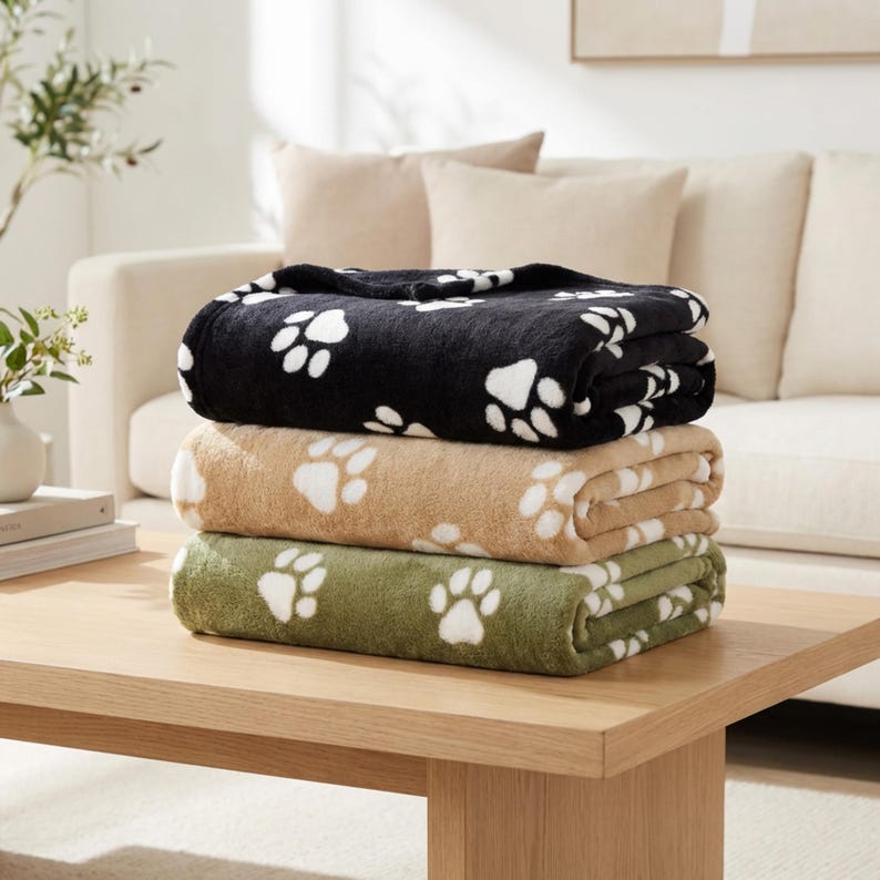 May include: A stack of three folded plush blankets, each featuring a repeating white paw print pattern. The top blanket is black, the middle blanket is tan, and the bottom blanket is olive green. The blankets have a soft, fuzzy texture.