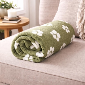 May include: A rolled-up olive green fleece blanket featuring a repeating pattern of white paw prints. The fabric has a soft, textured appearance, and the blanket is neatly coiled to display the interior and exterior layers of the patterned material.