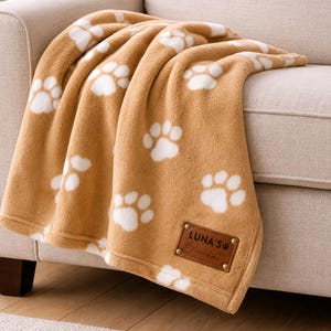 May include: A soft tan fleece blanket featuring a repeating white paw print pattern. The blanket includes a rectangular brown leather-like patch in the corner with gold metal rivets, embossed with the text LUNA'S Blanket and a small paw icon.