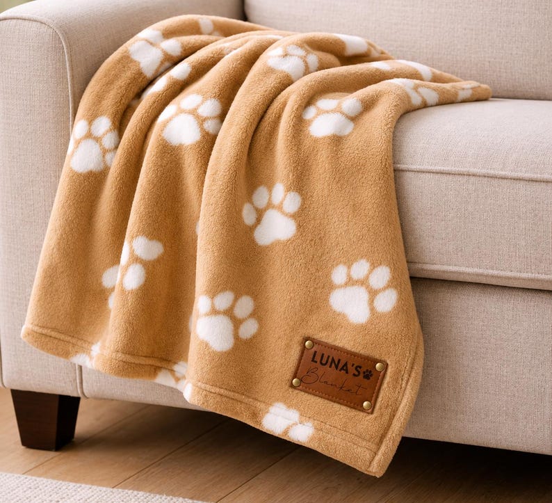 May include: A soft tan fleece blanket featuring a repeating white paw print pattern. The blanket includes a rectangular brown leather-like patch in the corner with gold metal rivets, embossed with the text LUNA'S Blanket and a small paw icon.
