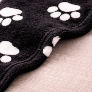 May include: A soft, black fleece blanket featuring a repeating pattern of white paw prints. The fabric has a thick, plush texture with a finished stitched edge that curves along the side, showcasing the dense, fuzzy material of the blanket.