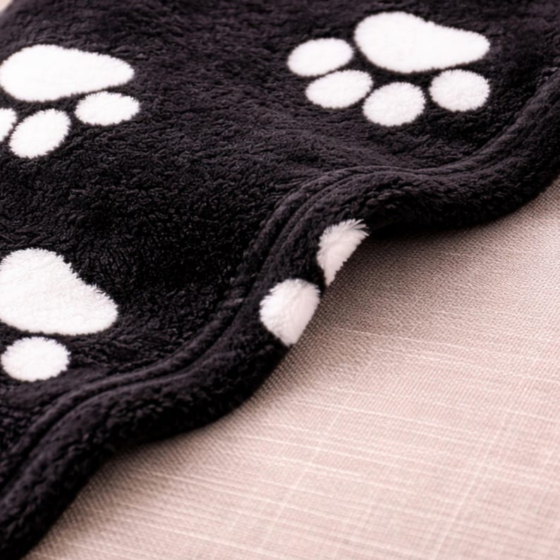 May include: A soft, black fleece blanket featuring a repeating pattern of white paw prints. The fabric has a thick, plush texture with a finished stitched edge that curves along the side, showcasing the dense, fuzzy material of the blanket.
