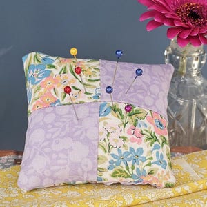 May include: A square pincushion made of patchwork fabric with a floral pattern. The pincushion is filled with stuffing and has several colorful pins sticking out of it. The pincushion is sitting on a yellow fabric surface.