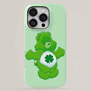 May include: A light green smartphone case featuring a graphic of a green bear character with a white belly badge displaying a dark green four-leaf clover. The bear has a small heart-shaped nose and is centred on the back of the phone case.