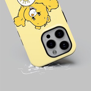 以下が含まれることがあります： 太陽の帽子をかぶった漫画のクマが描かれた黄色のiPhoneケース。ケースの背面にはカメラレンズの切り抜きがあります。