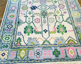 May include: A rectangular area rug featuring a cream-coloured field with a symmetrical floral and geometric pattern in shades of pink, green, and blue. The rug has a wide border with repeating motifs and a short white fringe on the ends.