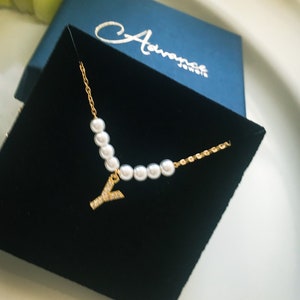 Puede incluir: Un collar de oro con una cadena de perlas y un colgante con la letra Y. El collar se muestra en una caja de terciopelo negro con una caja azul con el texto "Advance Jewelry" en la parte superior.