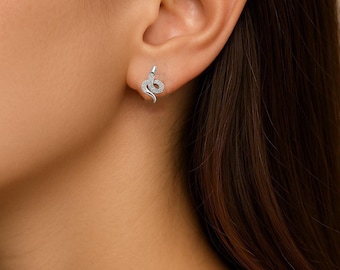 May include: A close-up of a silver snake stud earring. The earring is a detailed representation of a snake, with a coiled body and a raised head. The earring is set with small, sparkling stones.