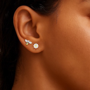 May include: Earrings with bee and daisy designs. The bee earring has a yellow and black striped body, silver head and wings. The daisy earring has white petals and a yellow centre. The earrings are worn on an ear.