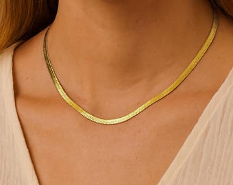 May include: A gold-colored herringbone chain necklace. The necklace is flat and lies smoothly against the skin. The chain is a bright, shiny gold color. The necklace is a classic and versatile piece of jewelry.