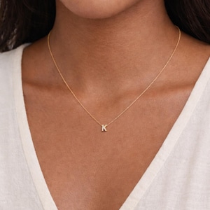 Può includere: Una delicata collana d'oro con un piccolo ciondolo con la lettera "K". Il ciondolo è appeso a una sottile catena. La persona indossa un top bianco. La collana è un design semplice ed elegante.