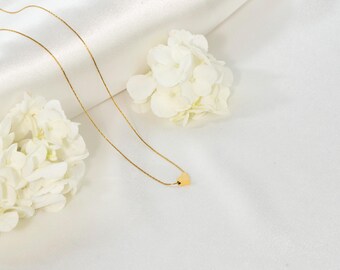 May include: A delicate gold chain necklace with a small heart pendant. The necklace is displayed on a white fabric background with white flowers.