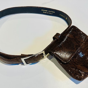 May include: A brown leather belt with a matching pouch. The belt is made of genuine leather and has a silver-colored buckle. The pouch is attached to the belt and has a textured, snake-like pattern.