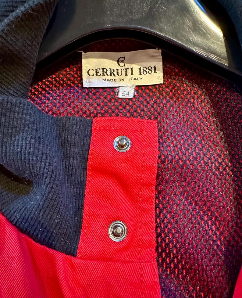 Puede incluir: Primer plano de una chaqueta roja y azul marino con la etiqueta "CERRUTI 1881 MADE IN ITALY". La chaqueta tiene un forro de malla y dos cierres de presi&oacute;n plateados. La etiqueta de talla dice "54".