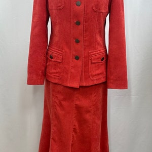 Könnte beinhalten: Ein roter Cordanzug mit Rock und Jacke. Die Jacke hat einen Stehkragen, sechs braune Knöpfe und zwei Pattentaschen. Der Rock ist A-förmig und reicht bis zu den Knien.