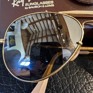 May include: Gold-framed Ray-Ban sunglasses with dark lenses, on a textured black surface. The Ray-Ban logo and "SUNGLASSES by BAUSCH & LOMB" are visible on the case. The lenses reflect the surrounding environment.