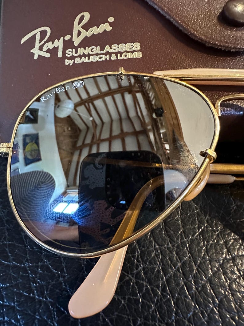May include: Gold-framed Ray-Ban sunglasses with dark lenses, on a textured black surface. The Ray-Ban logo and "SUNGLASSES by BAUSCH & LOMB" are visible on the case. The lenses reflect the surrounding environment.