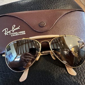 May include: Gold-framed Ray-Ban sunglasses with brown lenses and a brown leather case. The case is embossed with the Ray-Ban logo and the text "SUNGLASSES by BAUSCH & LOMB". The sunglasses have a classic aviator design.