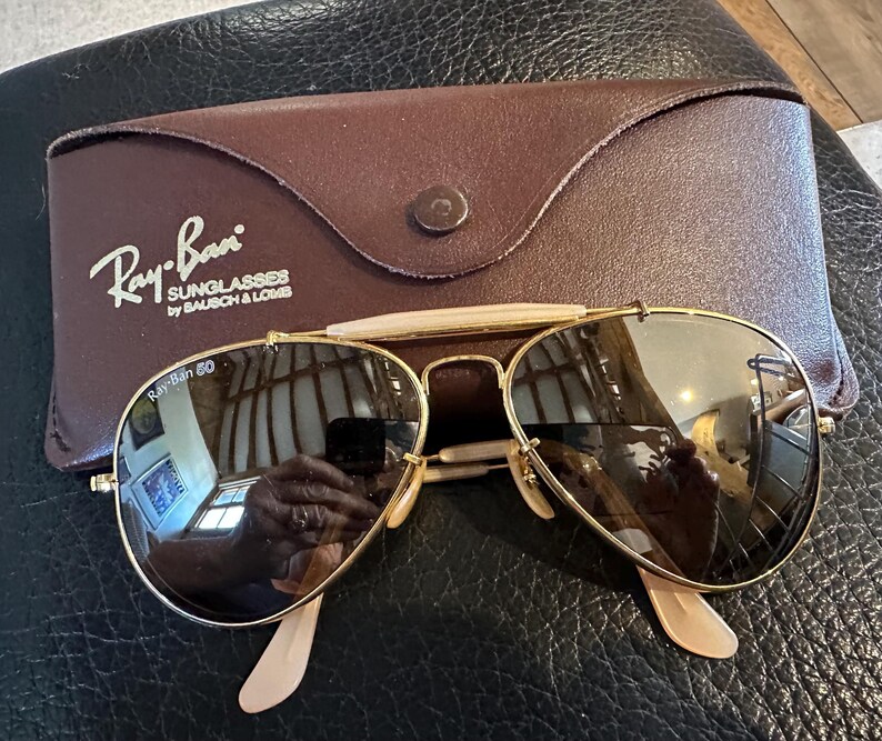 May include: Gold-framed Ray-Ban sunglasses with brown lenses and a brown leather case. The case is embossed with the Ray-Ban logo and the text "SUNGLASSES by BAUSCH & LOMB". The sunglasses have a classic aviator design.