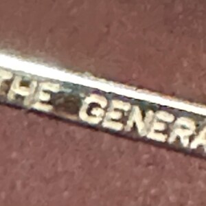May include: A close-up shot of a silver metal object with the words "THE GENERAL" engraved in white. The text is slightly blurred, but the letters are clearly visible against a dark background.