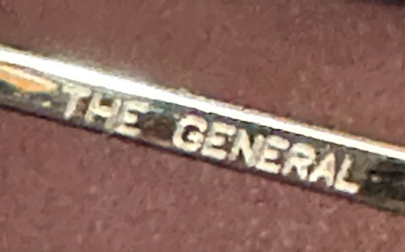 May include: A close-up shot of a silver metal object with the words "THE GENERAL" engraved in white. The text is slightly blurred, but the letters are clearly visible against a dark background.