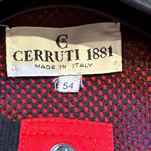 Puede incluir: Primer plano de una etiqueta de Cerruti 1881, fabricada en Italia. La etiqueta rectangular muestra el nombre de la marca y el logotipo en negro. El n&uacute;mero "54" est&aacute; impreso debajo. El fondo es una tela de puntos rojos y negros.