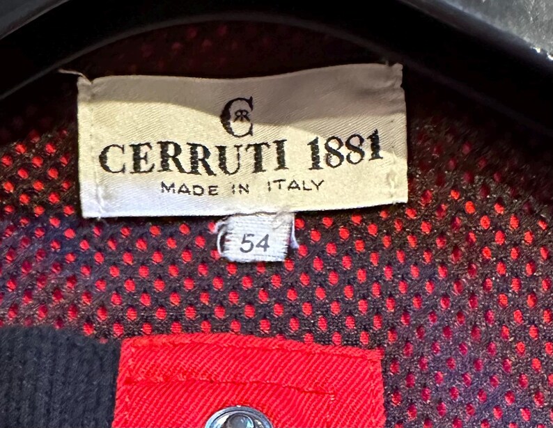 Puede incluir: Primer plano de una etiqueta de Cerruti 1881, fabricada en Italia. La etiqueta rectangular muestra el nombre de la marca y el logotipo en negro. El n&uacute;mero "54" est&aacute; impreso debajo. El fondo es una tela de puntos rojos y negros.