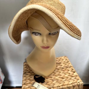 Puede incluir: Un sombrero de paja de ala ancha con un ribete beige claro y un interior beige. El sombrero se exhibe en una cabeza de maniquí. El sombrero tiene un diseño clásico, adecuado para la protección solar. El sombrero está hecho de paja tejida.