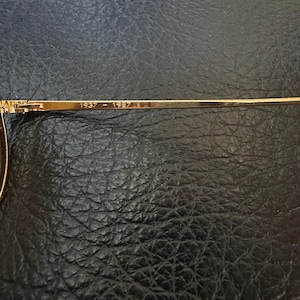 May include: Close-up of a gold-coloured eyeglass frame arm. The text "1937 - 1987" is printed on the arm. The frame is set against a dark, textured background.