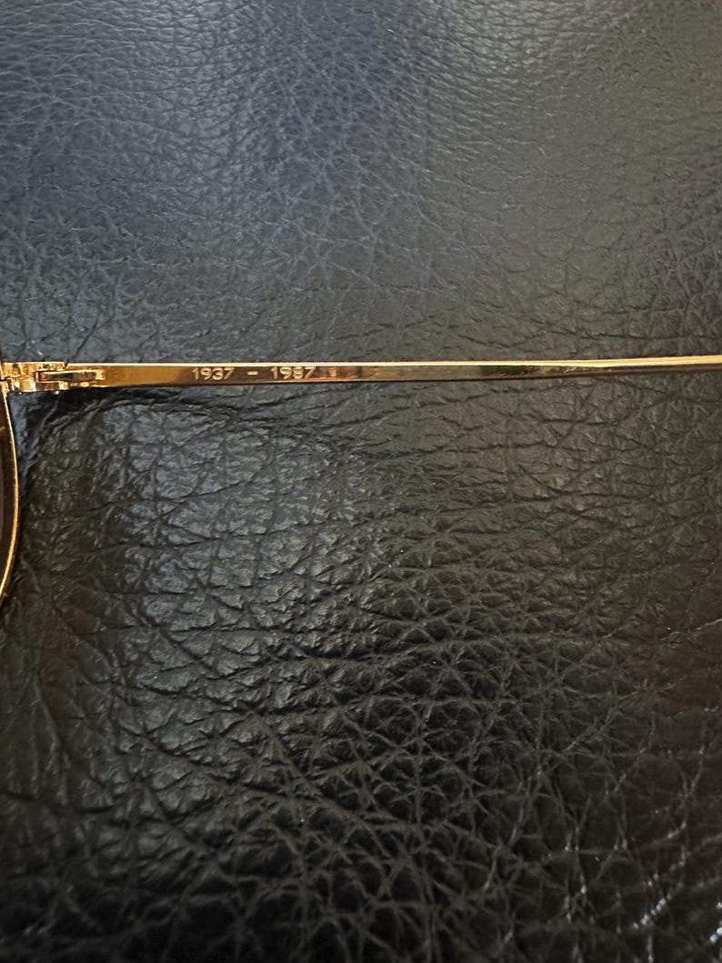 May include: Close-up of a gold-coloured eyeglass frame arm. The text "1937 - 1987" is printed on the arm. The frame is set against a dark, textured background.