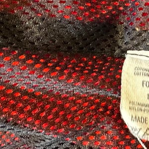 Puede incluir: Primer plano de una tela de malla negra y roja. Una etiqueta adjunta con texto en italiano, incluyendo "FODERA 100%" y "MADE IN ITALY". La tela tiene un dise&ntilde;o perforado.