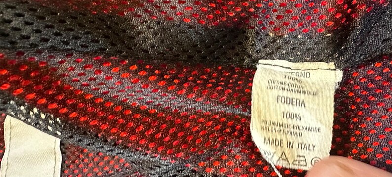 Puede incluir: Primer plano de una tela de malla negra y roja. Una etiqueta adjunta con texto en italiano, incluyendo "FODERA 100%" y "MADE IN ITALY". La tela tiene un dise&ntilde;o perforado.