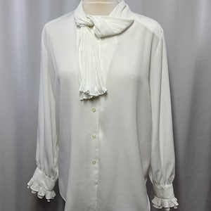 May include: A white long-sleeved blouse with a pleated bow tie at the neck and ruffled cuffs.