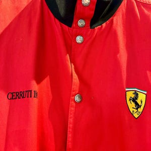 Puede incluir: Chaqueta roja con cuello negro y botones plateados. La chaqueta presenta el texto "CERRUTI 1881" en el lado izquierdo y un parche con el logotipo de Ferrari en el pecho derecho.