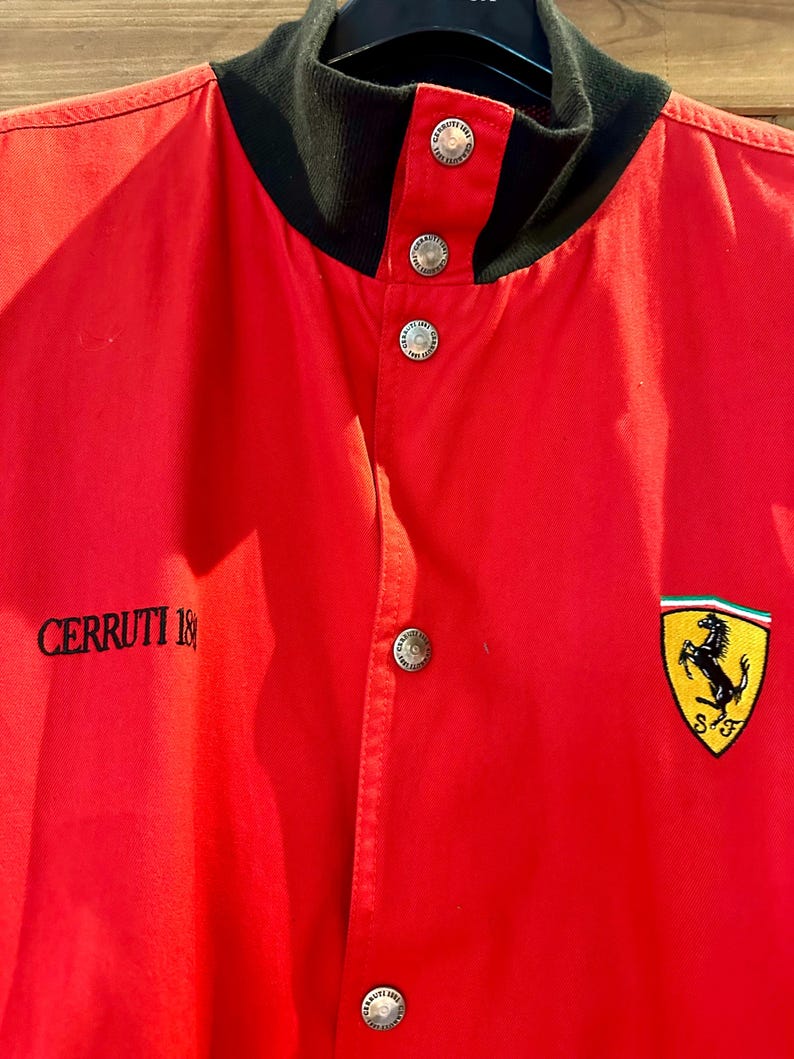 Puede incluir: Chaqueta roja con cuello negro y botones plateados. La chaqueta presenta el texto "CERRUTI 1881" en el lado izquierdo y un parche con el logotipo de Ferrari en el pecho derecho.