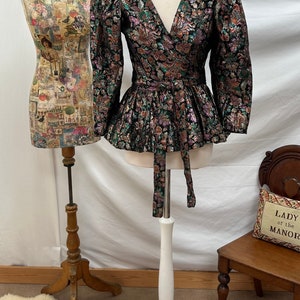 May include: A black, green, and gold brocade wrap top with a V-neckline and long sleeves. The top has a cinched waist and a ruffled hem. It is displayed on a white mannequin.