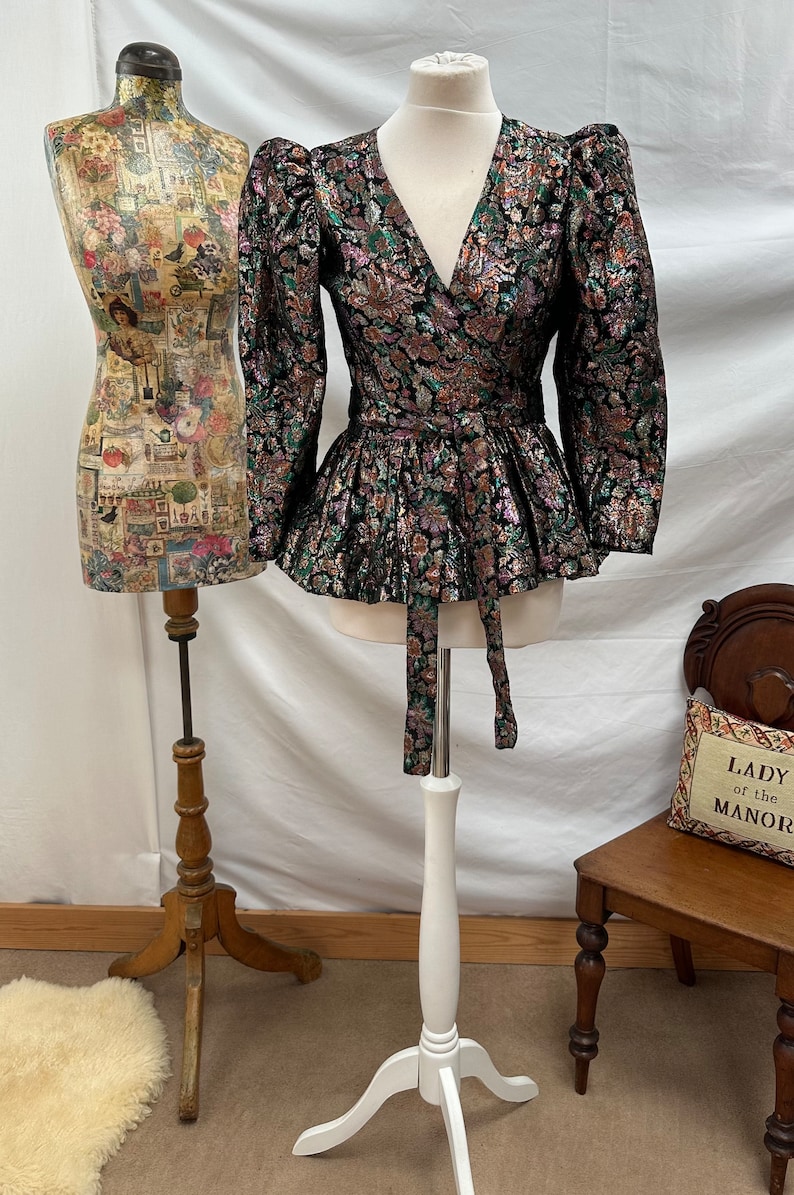 Puede incluir: Un top de envoltura de brocado negro, verde y dorado con cuello en V y mangas largas. El top tiene una cintura ajustada y un dobladillo con volantes. Se muestra en un maniqu&iacute; blanco.