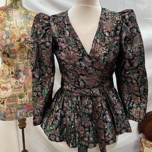 Puede incluir: Una blusa de gasa negra, verde y dorada con un dobladillo con volantes y un cintur&oacute;n de lazo. La blusa tiene un escote en V y mangas largas con hombros abullonados.