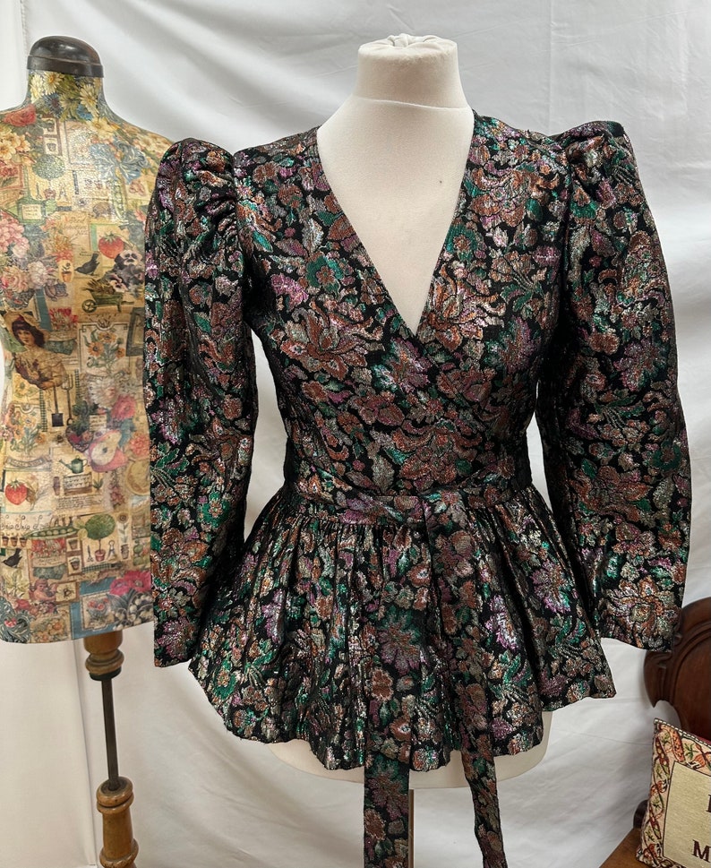 Puede incluir: Una blusa de gasa negra, verde y dorada con un dobladillo con volantes y un cintur&oacute;n de lazo. La blusa tiene un escote en V y mangas largas con hombros abullonados.